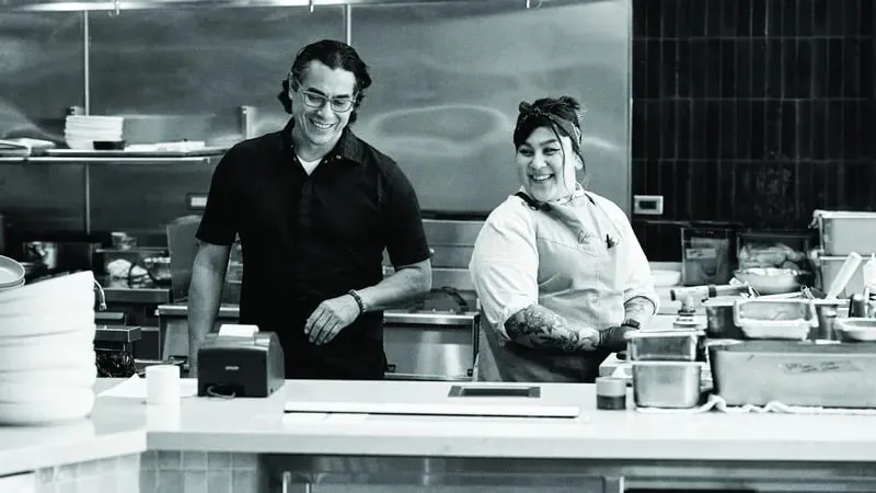 Chef Carlos Gaytan cooking with a fellow female chef in a kitchen